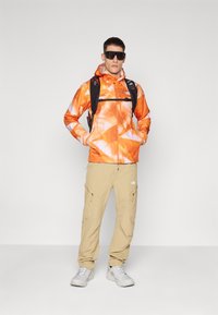 Lys orange jakke med et geometrisk mønster, beige cargosbukser og grå sneakers. Personen bærer mørke solbriller og en sort rygsæk.