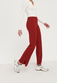 Pantalons rouges ajustés avec des fentes sur les côtés, en tissu doux. Associés à un haut ajusté à côtes blanc et des sneakers gris.