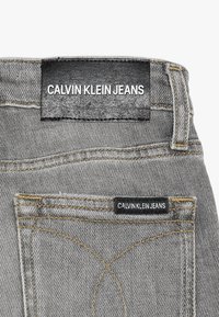 Harmaat farkut, joissa on musta nahkainen vyömerkki, johon on painettu valkoisella tekstillä "CALVIN KLEIN JEANS". Huomiota herättävä keltainen tikkaus.