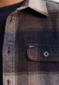Koszula flanelowa w jasnobrązowym i granatowym kratkę, z kołnierzykiem button-down, kieszonką na piersi oraz metką z logo Petrol blisko kieszonki.