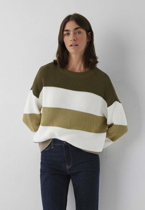 Femme aux cheveux bruns mi-longs portant un pull ample à rayures olive, blanc et beige, associé à un jean bleu foncé, debout contre un mur.