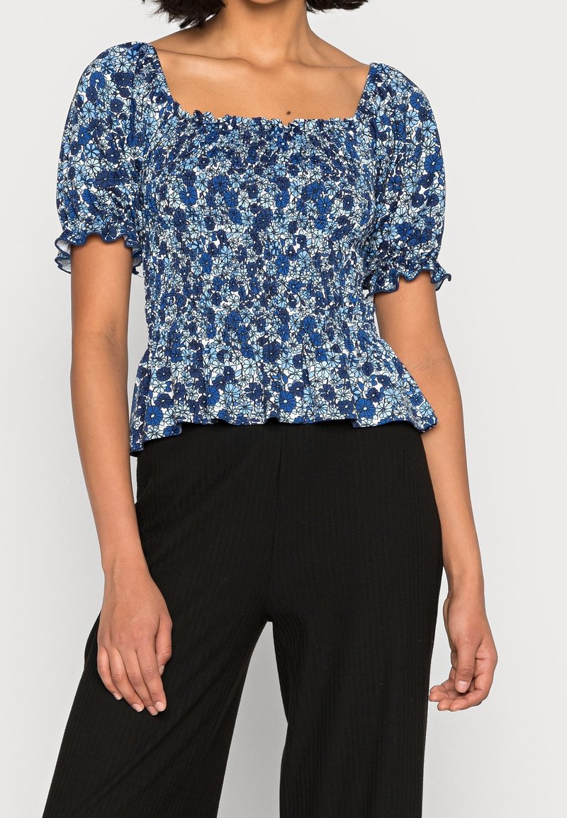 Blå blommig peplumtop med korta puffärmar, fyrkantig halsringning och smockdetaljer vid bysten; kombinerad med svarta byxor.