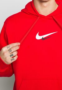 Röd hoodie av bomull med en frampocket, med en vit Nike-logotyp och en mindre svart swoosh-accent. Dragsko vid huvan.