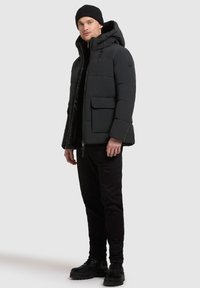 Schwarze gepolsterte Jacke mit einem Reißverschluss, Kapuze und Taschen vorne. Getragen mit schwarzen Hosen und schwarzen Schnürstiefeln. Glatte Textur und strukturiertes Design.
