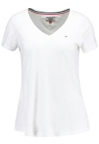 Camiseta blanca de cuello en V hecha de algodón con mangas cortas. Presenta un logo sutil y un detalle de rayas en negro, rojo y blanco en el cuello.