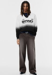 Personne avec des dreadlocks portant un pull dégradé noir et blanc avec du texte, un pantalon gris à jambes larges et des chaussures noires pointues sur un fond blanc.