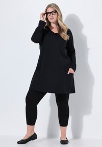 Schwarze Tunika mit langen Ärmeln und seitlichen Taschen, kombiniert mit schwarzen Leggings und strukturierten flachen Schuhen. Einfach und figurnah geschnitten, mit einem V-Ausschnitt.