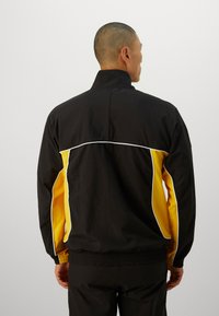 Sort og gul letvægtsjakke med ståkrave, elastisk talje og hvid piping langs skuldersømmene.