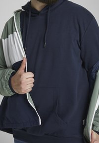 Felpa con cappuccio blu marina con tasca frontale, abbinata a una giacca verde e bianca con zip, con polsini elastici e vestibilità rilassata.
