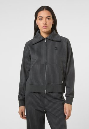 Jeune femme portant une veste zippée à fines rayures gris foncé et un pantalon assorti, debout devant un fond clair uni, face à la caméra.