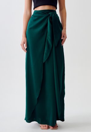 Kvinde iført en mørkegrøn omslagsnederdel bundet i taljen, kombineret med nude åbne sandaler og en sort kort top mod en ensfarvet baggrund.