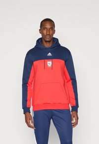Sweatshirt com capuz em mistura de poliéster vermelho e azul-marinho, com um bolso canguru e logótipo da equipe no peito. Mangas longas com cores contrastantes.