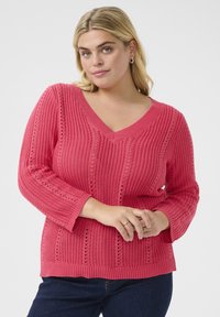Pull en tricot rose avec décolleté en V, présentant un tissu texturé, des motifs complexes de couture ouverte et des manches trois-quarts.