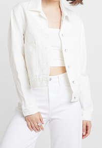 Biała, skrócona kurtka dżinsowa z kołnierzykiem, z guzikami z przodu i dwiema kieszeniami na piersi, zestawiona z białym prążkowanym topem i spodniami o wysokim stanie.