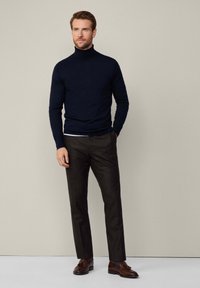 Marinblått polotröja i ull tillsammans med mörka strukturerade byxor och bruna loafers. Modellen står mot en neutral bakgrund med händerna längs sidorna.