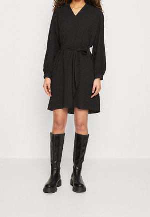 Woman wearing a knee-length black dress with long sleeves, a waist tie, and black knee-high chunky boots, standing against a white background.