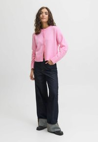 Roza pletena jopica s krožnim ovratnikom, dolgimi rokavi in gumbom, v kombinaciji s temno širokimi jeans hlačami in debelimi črnimi čevlji.