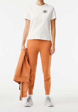Person som bär en vit kortärmad Lacoste-t-shirt, orangea sweatpants, vita sneakers och håller en matchande orange jacka i vänster hand.