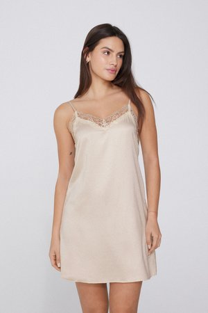 Femme aux longs cheveux foncés portant une robe nuisette en satin beige avec bordure en dentelle, debout devant un fond clair uni.