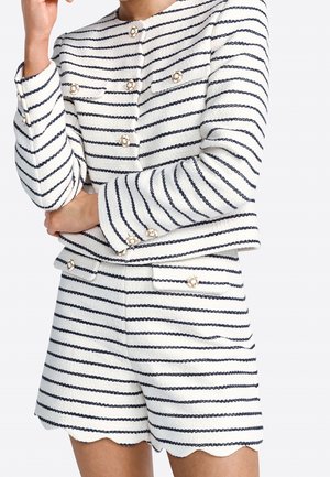 Vrouw draagt een wit pak van een getextureerde stof met zwarte horizontale strepen, gouden knopen, lange mouwen en korte broek met geschulpte zoom.