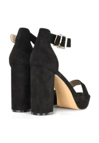 Sandalias de tacón alto de ante negro con tacones gruesos en bloque, correa al tobillo con hebilla y diseño de punta abierta; textura suave y detalles mínimos.