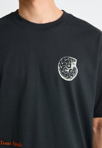 Sort t-shirt med et lille hvidt fossil grafik og "Texas Fossils" broderet i orange nær sømmen, iført af en person.