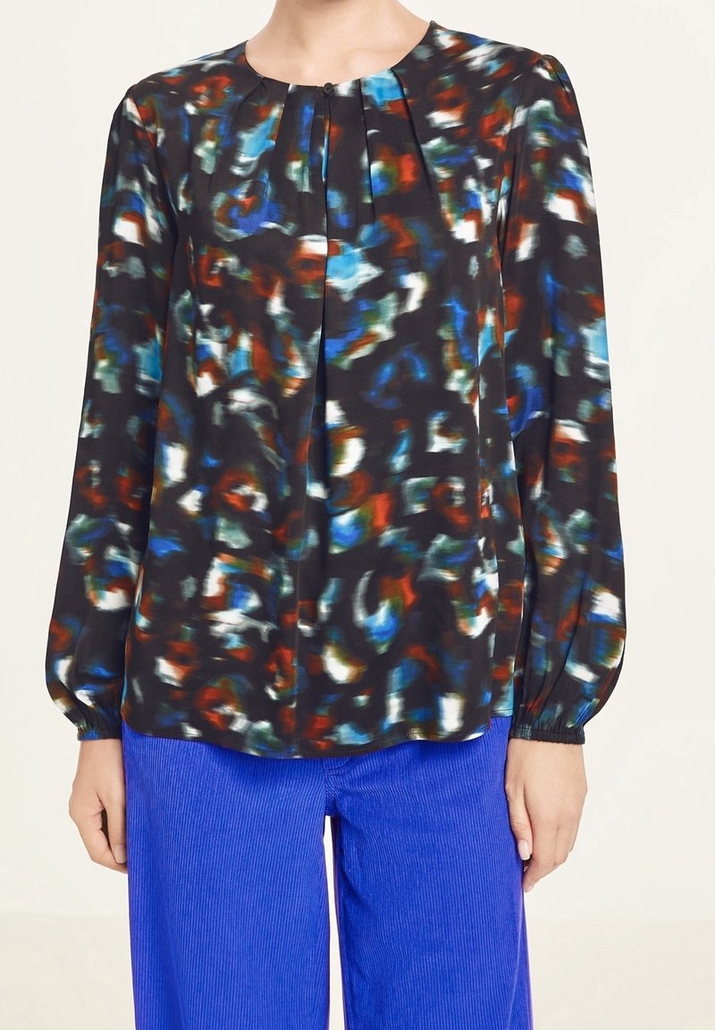 Bluzka z długim rękawem z abstrakcyjnym, wielokolorowym wzorem w kolorach czarnym, niebieskim, czerwonym i białym, zestawiona z jaskrawo niebieskimi spodniami.