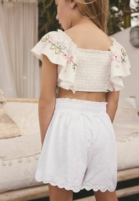 Top court smock blanc avec des manches à volants et broderies florales, associé à un short blanc avec un ourlet festonné et une texture florale.
