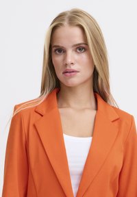Blazer arancione con colletto a rever, spalle strutturate e tessuto liscio. Indossato sopra una maglietta bianca, mette in evidenza linee pulite e vestibilità.