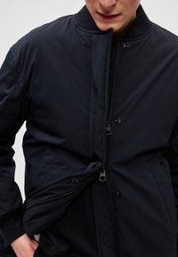 Námornícka modrá bomber bunda so zipsom a zapínaním na gombíky, vybavená bočnými vreckami a prešívanou vnútornou podšívkou pre zvýšenie tepla.