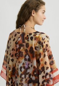 Cappa in chiffon con stampa leopardata marrone e arancione, texture trasparente e nodo al collo. Include un'accennata decorativa sul tessuto.