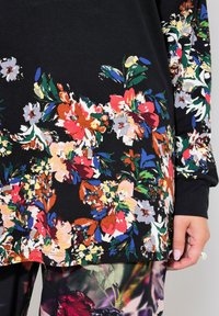 Schwarzes Oberteil mit floralem Muster, das lebhafte rote, orange und blaue Blumen zeigt. Aus weichem Stoff und lockerer Passform mit langen Ärmeln, die filigrane Details zur Schau stellen.