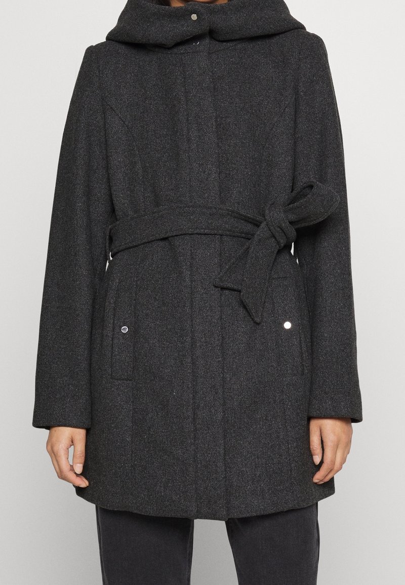 Manteau en laine gris foncé avec ceinture à la taille nouée en nœud et poches à rabat avec fermeture pression, porté sur un pantalon noir.