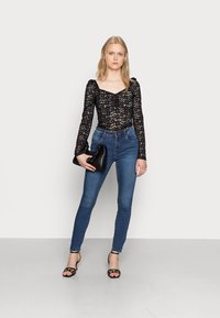 Jovem mulher vestindo uma blusa de renda preta, jeans skinny azuis, saltos altos pretos com tiras, segurando uma carteira preta franzida contra um fundo branco.