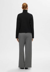Črni pleten turtleneck pulover s sproščenim krojem, v kombinaciji s sivimi širokimi hlačami in črnimi čevlji, gledano s hrbta.
