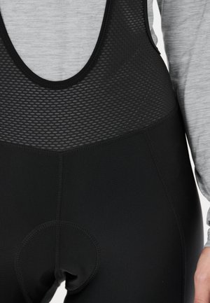 Close-up van zwarte gevoerde fietsbibshorts gedragen over een grijze top met lange mouwen, met een zichtbare mesh bovenstuk en gedetailleerde stiksels.