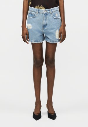 Osoba w jasnoniebieskich, postarzanych szortach dżinsowych i czarnych szpilkach z ostrymi noskami, stojąca na tle jednolitego, jasnego tła.