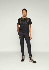 Crna majica s zlatnim detaljima i teksturiranim uzorkom, uparena s crnim hlačama s uzorkom i crno-zlatnim čizmama. Minimalistički sportski stil.