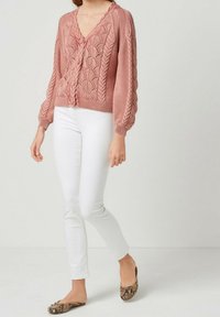 Donna che indossa un cardigan rosa lavorato a maglia con bottoni e motivi a foglia, pantaloni bianchi aderenti e scarpe slip-on con motivo in pelle di serpente.