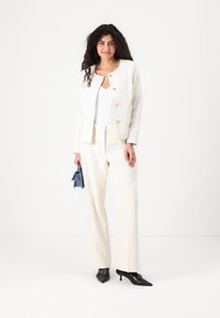 Vit texturerad blazer med guldfärgad knappar, kombinerad med en vit topp, beige byxor med ränder, svarta spetsiga klackar och en liten blå kuvertväska.