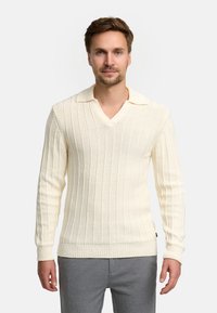 Maglione a maglia color crema con scollo a V, texture a coste verticali, maniche lunghe e colletto ripiegato. Indossato con pantaloni grigi.