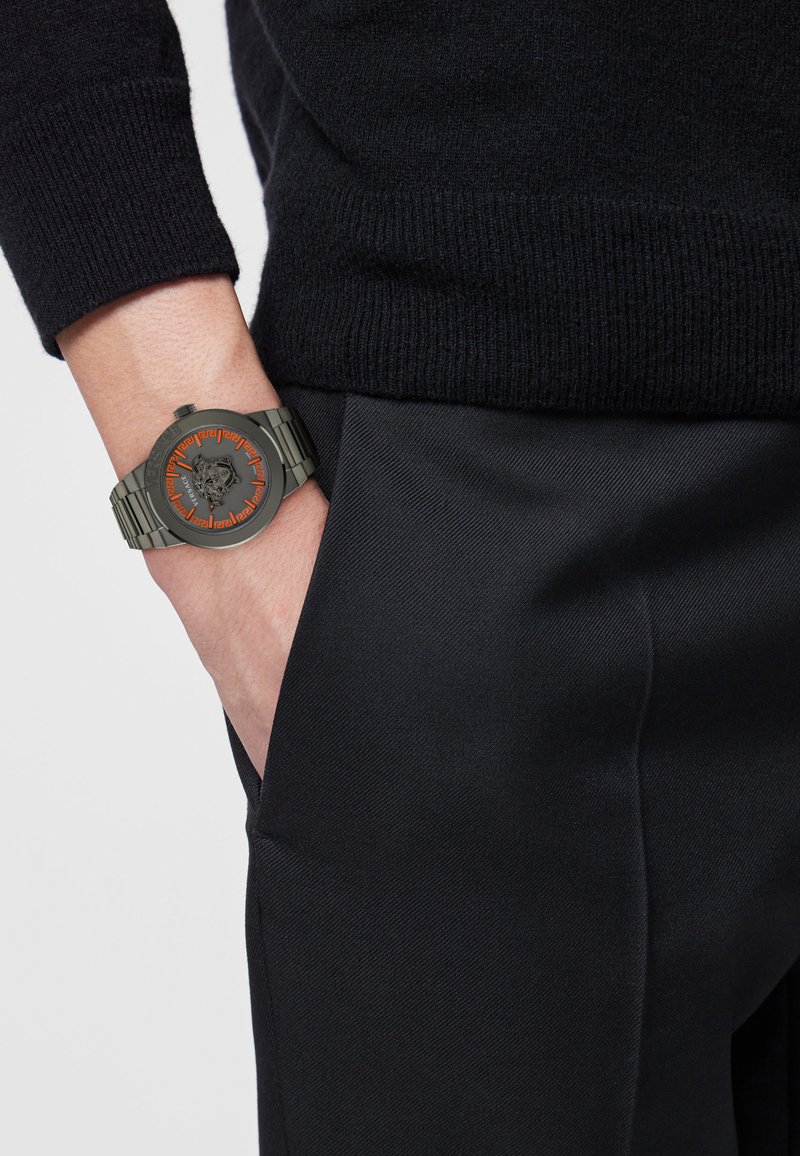 Orologio con un quadrante tondo di colore grigio scuro, decorato con un design in rilievo e dettagli arancioni, attaccato a un bracciale metallico di colore argento. Indossato su un abbigliamento nero.