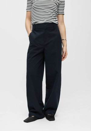 Personne portant un pantalon noir à jambes larges, des chaussures noires pointues et un haut à manches courtes rayé noir et blanc, debout avec les mains dans les poches.