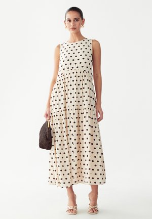 Frau trägt ärmelloses cremefarbenes Maxikleid mit schwarzen Punkten, hält eine geflochtene braune Handtasche und trägt beige Sandalen.