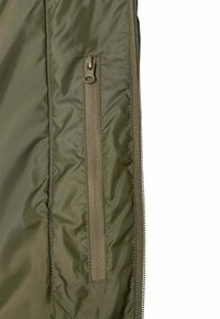 Vue latérale d'une veste vert olive avec une poche verticale zippée et une bande de fermeture éclair visible sur le bord droit.