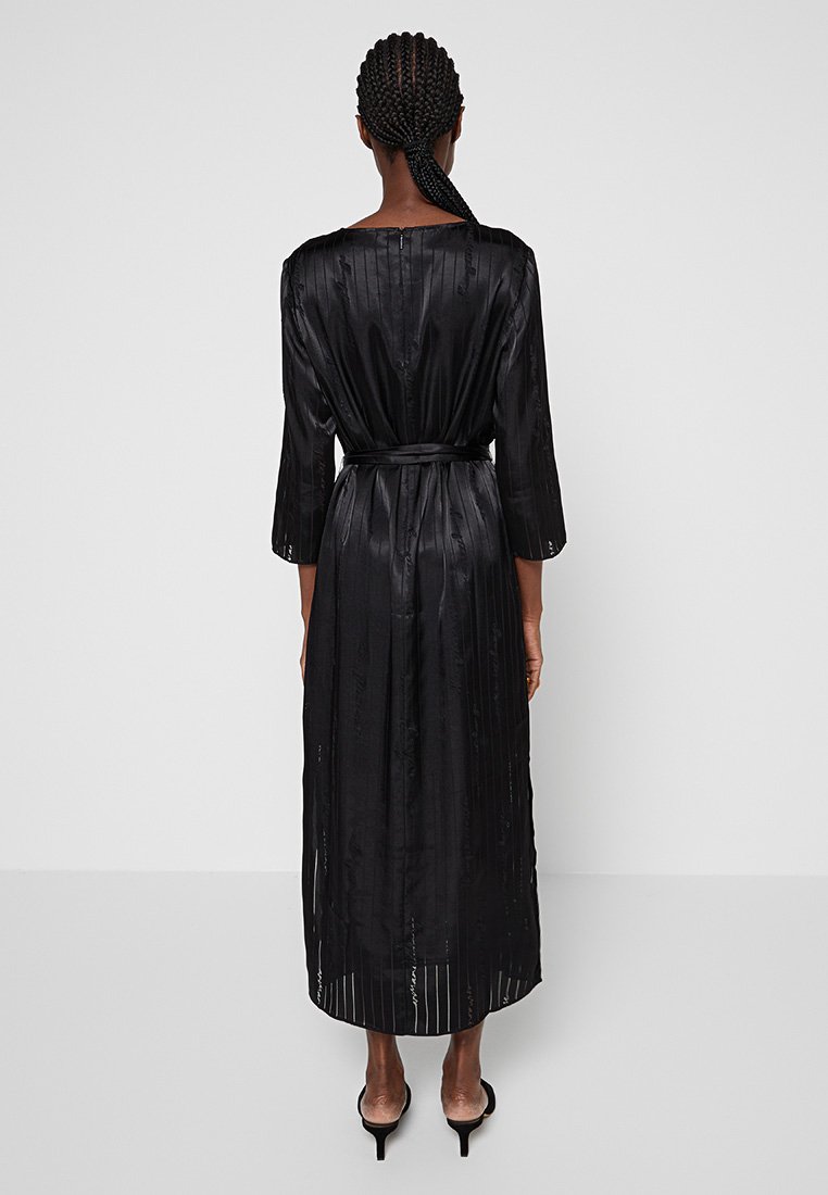 Dlouhá černá saténová šaty se třičtvrtinovými rukávy, pasem se sponou a vertikálními pruhy pro texturu. Délka po střed lýtek, s uvolněným střihem.