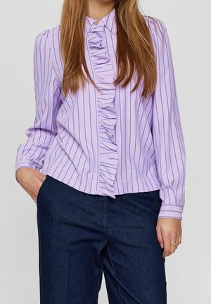 Kobieta ubrana w fioletową bluzkę z długim rękawem z niebieskimi pionowymi paskami i falbankami, w połączeniu z ciemnoniebieskimi spodniami z wysokim stanem.