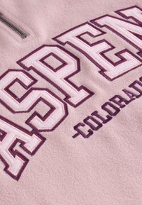 Mjuk rosa fleece-tyg med stora broderade bokstäver som bildar "Aspen" och mindre text "Colorado" i mörklila tråd.