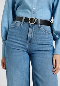 Cinturón de cuero negro con una hebilla circular de plata, usado sobre una camisa de denim azul claro y combinado con unos vaqueros de cintura alta azul.