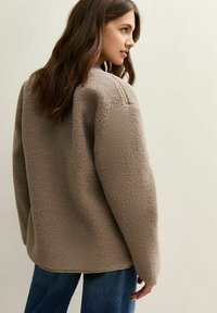 Beige fleecjas met een gestructureerd oppervlak, oversized pasvorm en verlaagde schouders, gecombineerd met blauwe spijkerbroeken. Gezien vanachter.